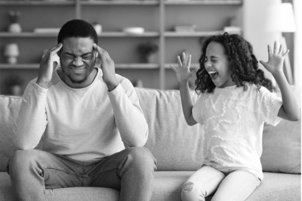 Ab wann ärgern Kinder bewusst? Tochter lärmt auf dem Sofa, während der Vater daneben sitzt und sich die Ohren zuhält – Alltagsszene bei „Kind provoziert“